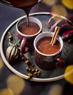 Hot chocolate pours into a mug next to another with a cinnamon stick. Spices are scattered around the table.