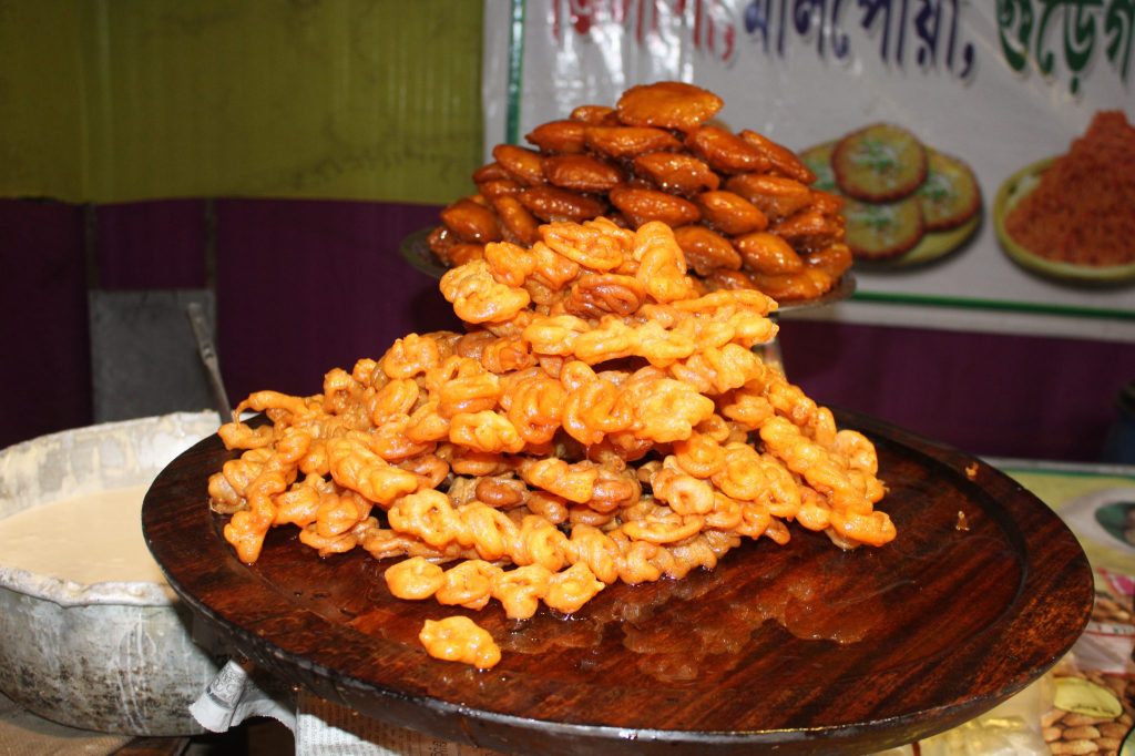A close-up view of piles of jalebi sweets arranged on a wooden tray, with a metal pot containing batter nearby, displayed at a food stall.